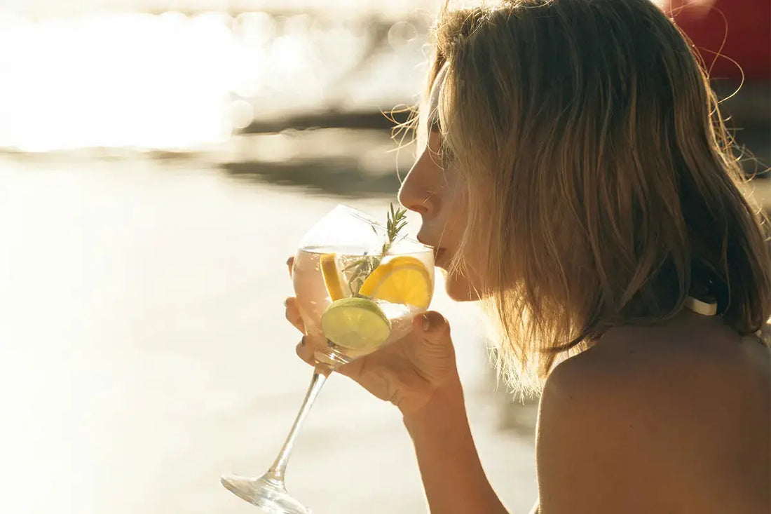 Femme sirotant un gin tonic avec tranche de citron, en apéritif au coucher du soleil, dans un verre à cocktail ballon.