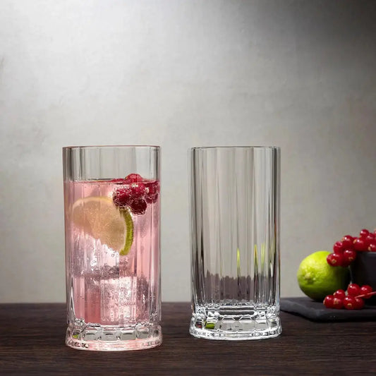 Deux verres à cocktail highball en cristal facetté, dont l’un rempli d’un cocktail pétillant rose avec citron vert et groseilles. Élégance et raffinement pour vos boissons longues.
