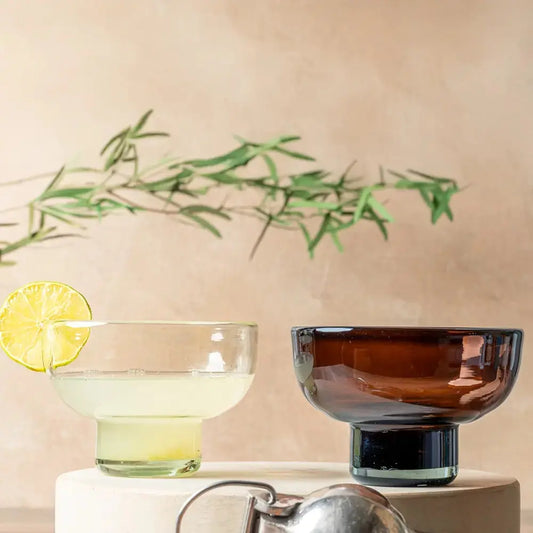 Set de deux verres à mezcal en verre soufflé, l’un transparent avec un cocktail décoré d’une rondelle de citron vert, l’autre en verre ambré. Présentés sur un socle beige avec une branche d’olivier en arrière-plan, créant une ambiance méditerranéenne et raffinée.
