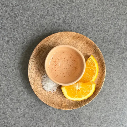 Vue de dessus d'un verre à mezcal traditionnel en céramique beige, posé sur une assiette en bois avec des tranches d'orange et du sel de mer. Une touche de piment est saupoudrée à la surface du liquide, mettant en avant un rituel de dégustation mexicain authentique.