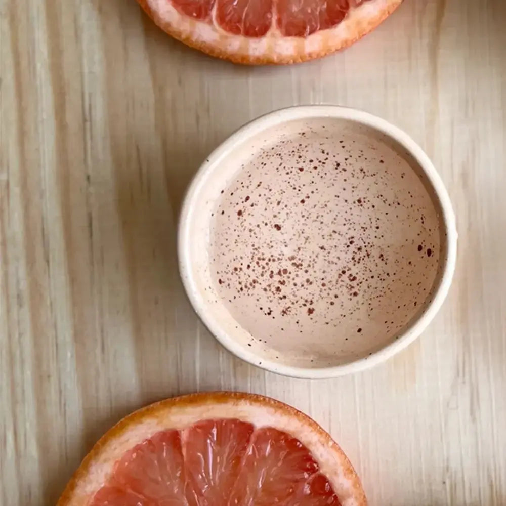 Verre à mezcal traditionnel en céramique beige, vu de dessus, avec une texture mouchetée caractéristique. Il est posé sur une planche en bois claire, accompagné de tranches de pamplemousse, évoquant une dégustation authentique et artisanale.