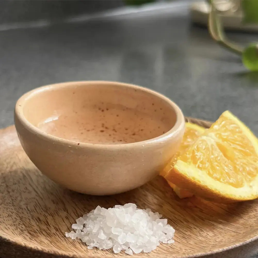 Petit verre à mezcal traditionnel en céramique beige, posé sur une assiette en bois. À côté, des tranches d'orange juteuses et une petite pile de sel de mer, évoquant le rituel de dégustation du mezcal. L'arrière-plan flou met en valeur l'esthétique artisanale et authentique de la scène.