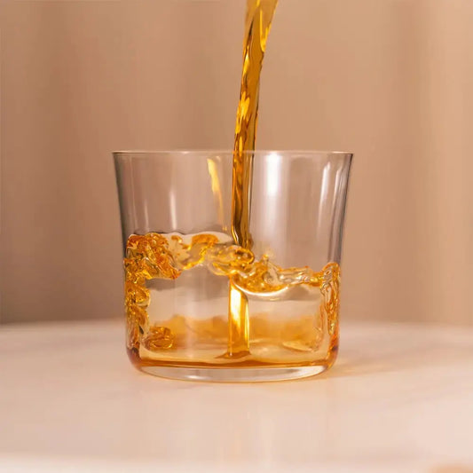 Versement d’un spiritueux ambré dans un verre à cocktail Lowball, posé sur une surface claire avec un fond beige.