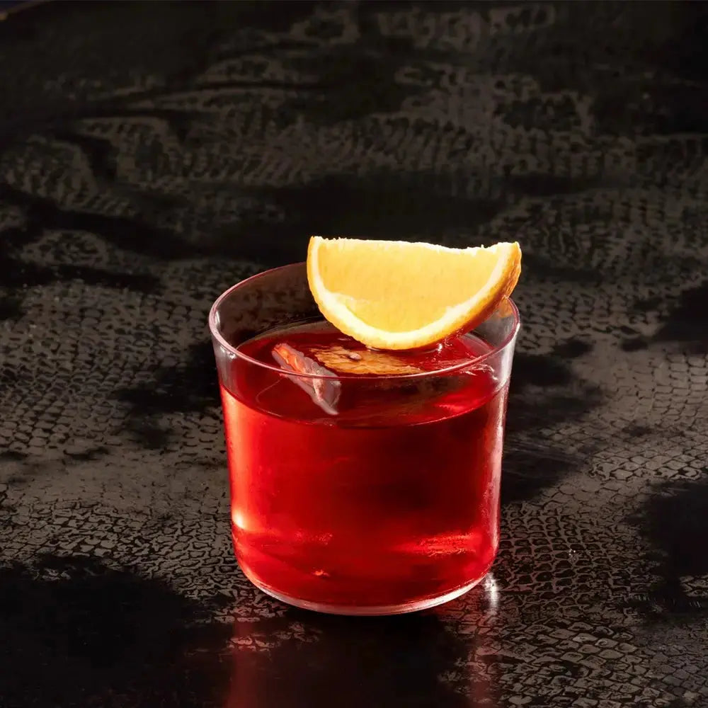 Verre à cocktail Lowball rempli d’un Negroni, surmonté d’une tranche d’orange, posé sur une table sombre texturée.