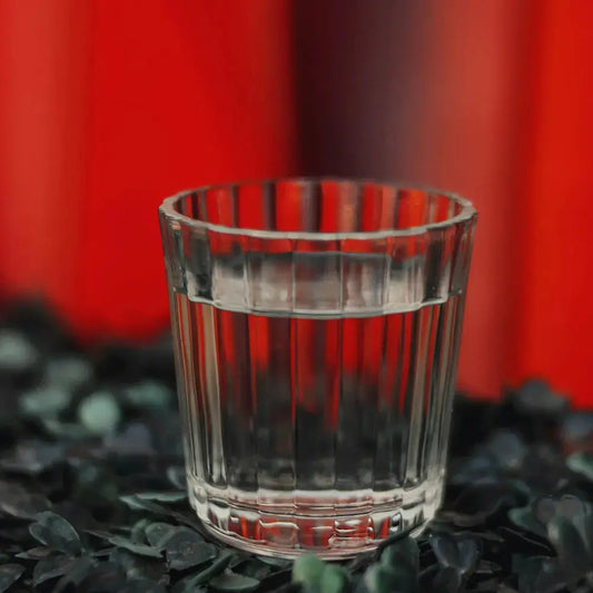 Verre à mezcal Veladora en cristal, rempli de liquide transparent, posé sur un lit de feuilles sombres avec un fond rouge vibrant.