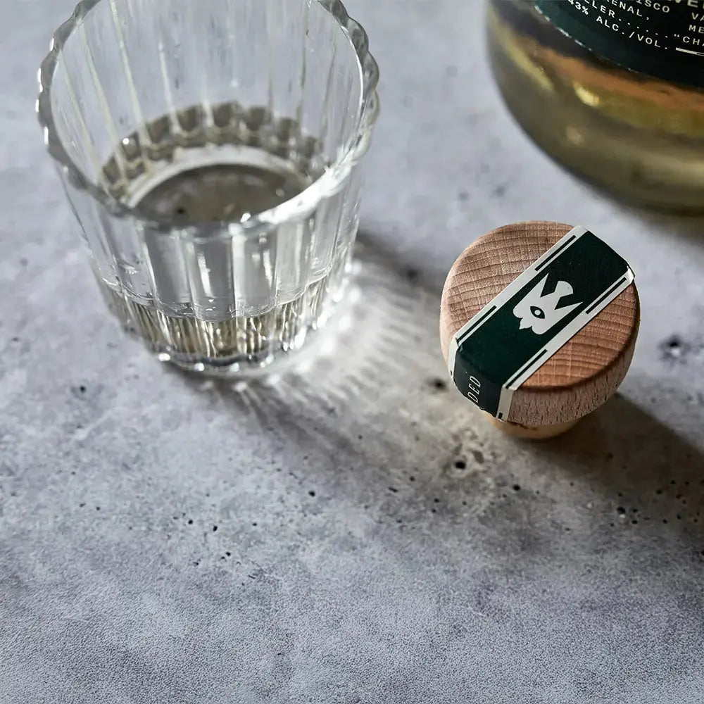 Verre à mezcal de style veladora en cristal strié, partiellement rempli de mezcal, posé sur une surface en béton brut. À côté, un bouchon en bois avec un sceau vert foncé représentant un oiseau, et en arrière-plan, une bouteille de mezcal légèrement floutée.