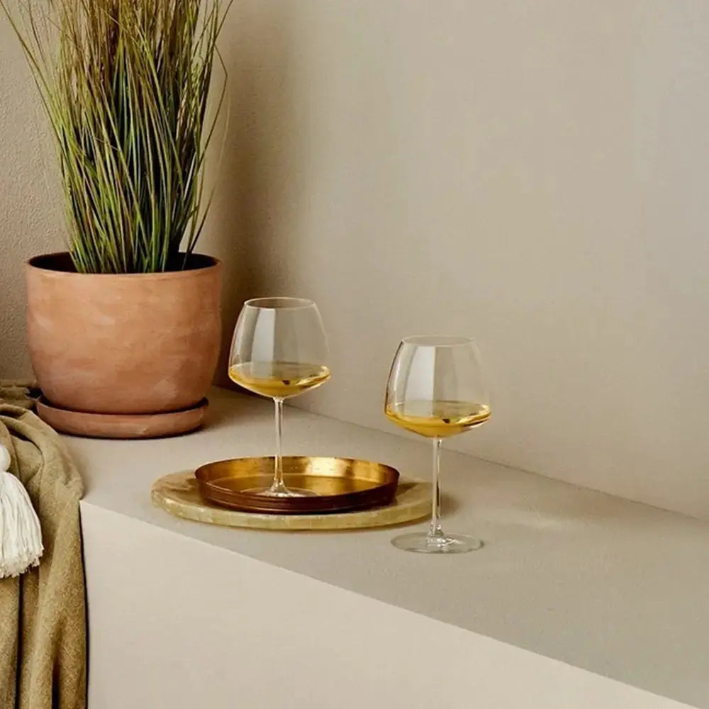 Deux verres à vin Chardonnay élégants, remplis de vin blanc doré, posés sur un plateau doré dans un décor chaleureux avec une plante en pot et une ambiance tamisée.