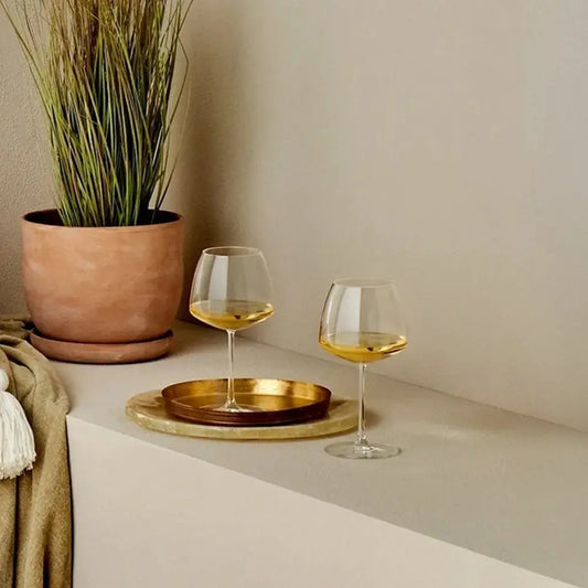 Deux verres à vin Chardonnay élégants, remplis de vin blanc doré, posés sur un plateau doré dans un décor chaleureux avec une plante en pot et une ambiance tamisée.