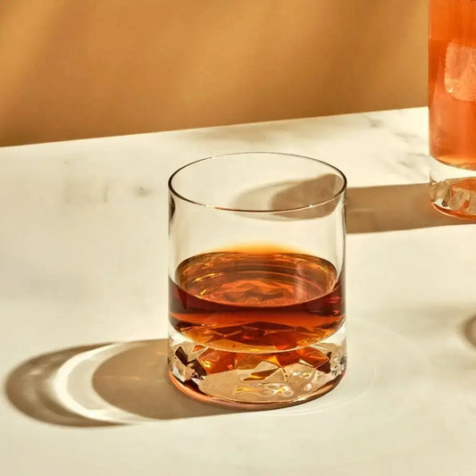 Verre à whisky effet ondulé transparent rempli, posé sur une table en marbre clair avec lumière chaude et ombres marquées.