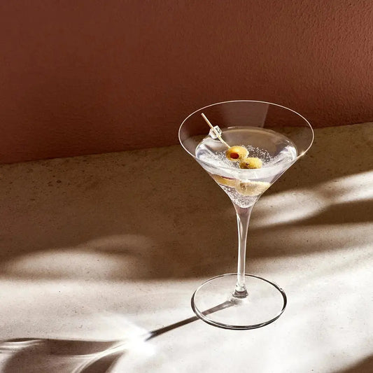 Verre à Martini vintage en cristal transparent, rempli d'un cocktail avec des olives en garniture, posé sur une table en pierre avec une lumière douce et une ombre élégante.