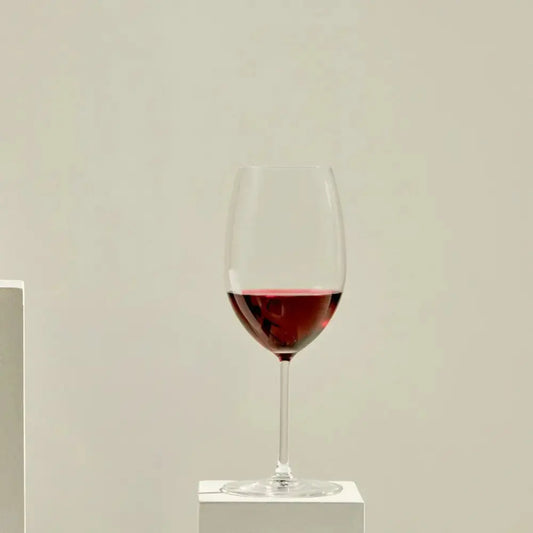 Verre à vin Bordeaux rempli de vin rouge, posé sur un socle blanc, avec un fond beige minimaliste.
