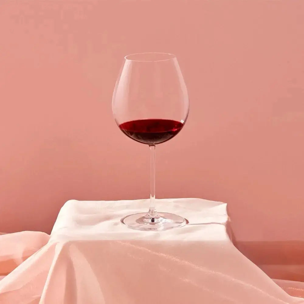 Verre à vin Bourgogne élégant, rempli de vin rouge, posé sur un tissu drapé dans une ambiance douce aux teintes rosées.