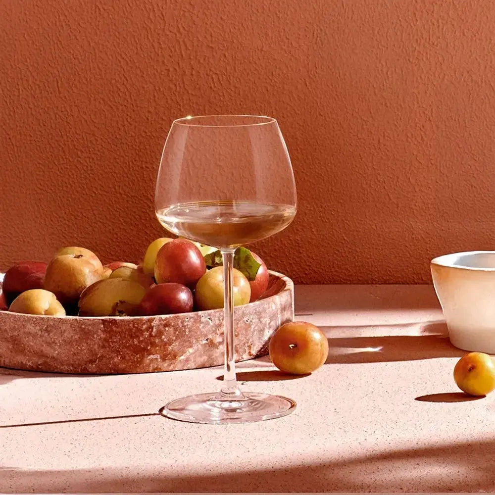 Verre à vin Chardonnay rempli de vin blanc, posé sur une table ensoleillée avec une coupe de fruits, contre un mur texturé de couleur terracotta.