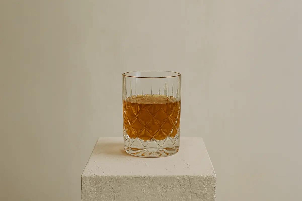 Verre à whisky en cristal, rempli d’un spiritueux ambré, posé sur un socle texturé beige dans une ambiance sobre et élégante.