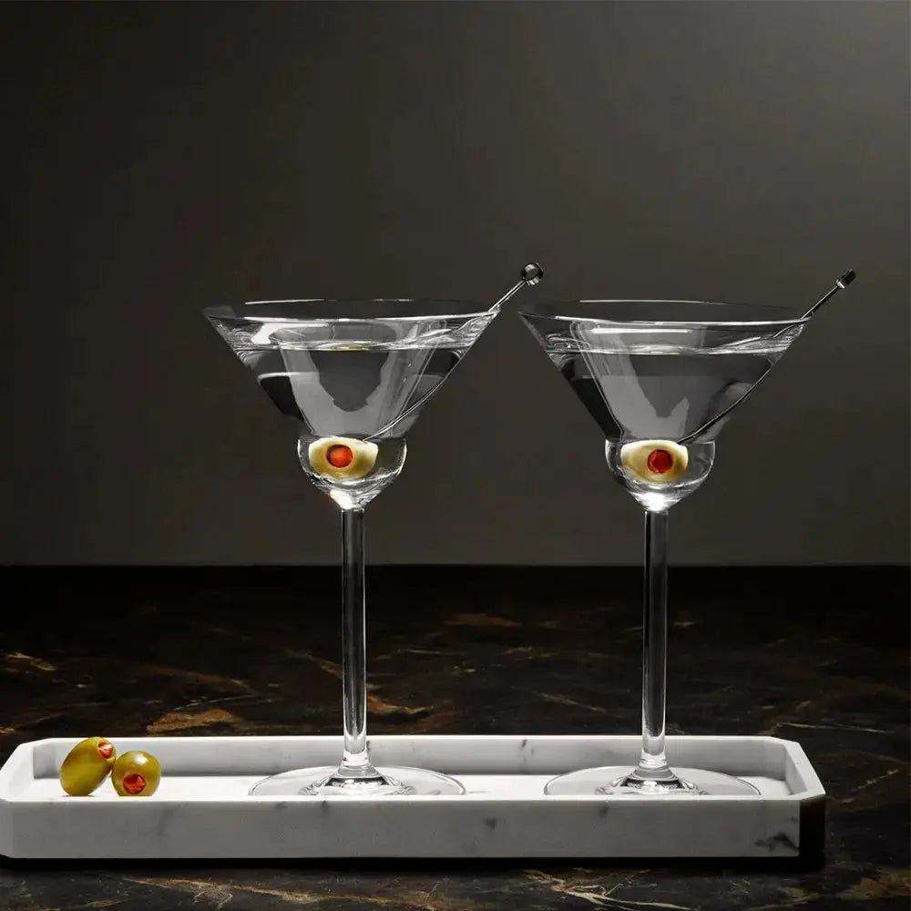 Ensemble de 2 verres pour Martini arrondis sur une planche en marbre blanc. Les deux verres sont remplis de dry martini. 