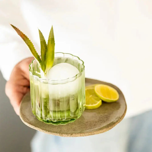 Verre à cocktail lowball en verre vert texturé, servi avec un cocktail rafraîchissant, un grand glaçon sphérique et une garniture de feuilles d’ananas. Présenté sur un plateau en céramique avec des quartiers de citron vert pour une ambiance élégante et raffinée.