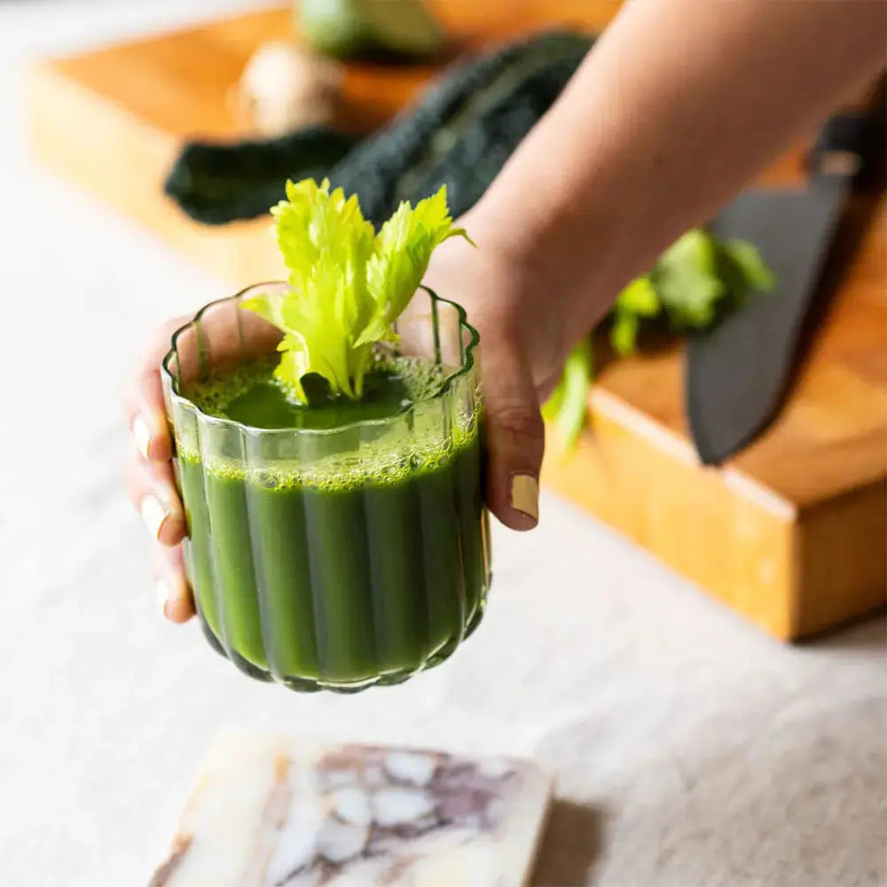 Verre à cocktail lowball en verre vert translucide, tenu à la main et rempli d’un cocktail vert décoré d’une branche de céleri. Une mise en scène élégante et naturelle, idéale pour déguster des boissons fraîches et originales.