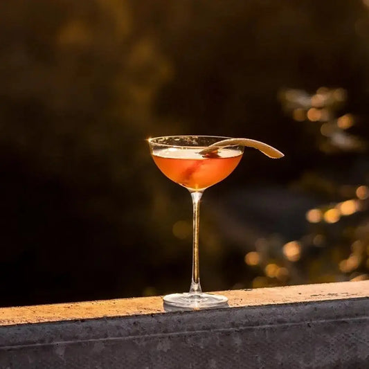 Coupe à cocktail Hepburn remplie d’un cocktail ambré, garnie d’un zeste d’agrume, capturant l’ambiance chaleureuse d’un coucher de soleil.