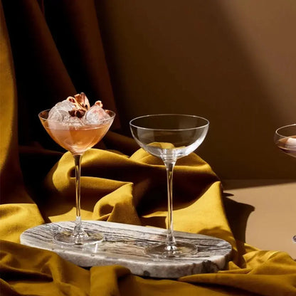 Ensemble de coupes à cocktail Hepburn, élégamment disposées sur un tissu doré, avec une coupe remplie d’un cocktail glacé, capturant une ambiance sophistiquée et chaleureuse.