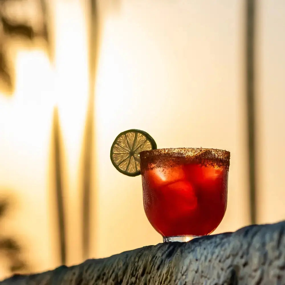 Verre à cocktail rempli d’une Michelada rouge intense, avec un bord salé et épicé, garni d’une rondelle de citron vert. Capturé au coucher du soleil pour une ambiance mexicaine authentique.