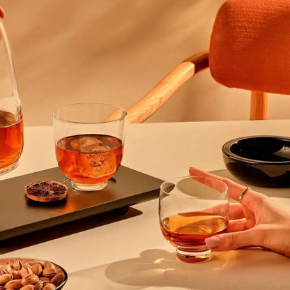 Mains élégantes tenant un verre à cocktail bas rempli d’un spiritueux ambré. Une ambiance chaleureuse avec des verres à whisky sur une table raffinée, accompagnés d’agrumes séchés et de pistaches.