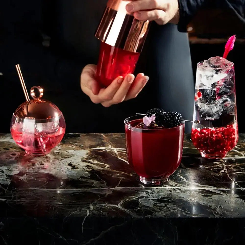 Mixologue en action préparant un cocktail raffiné. Trois verres à cocktail design, garnis de fruits rouges et de glaçons, posés sur un comptoir en marbre. Une scène sophistiquée évoquant l’art de la mixologie.