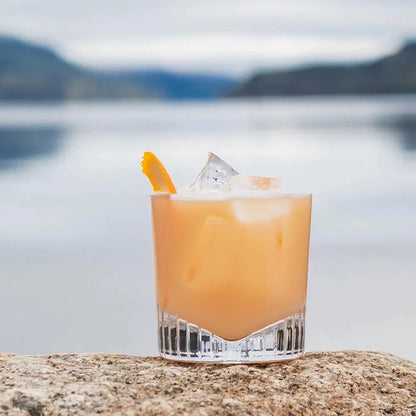 Set de 4 verres de whisky Caldera rempli d'un cocktail frais. Le verre est posé sur une roche et au fond il y a un lac. 