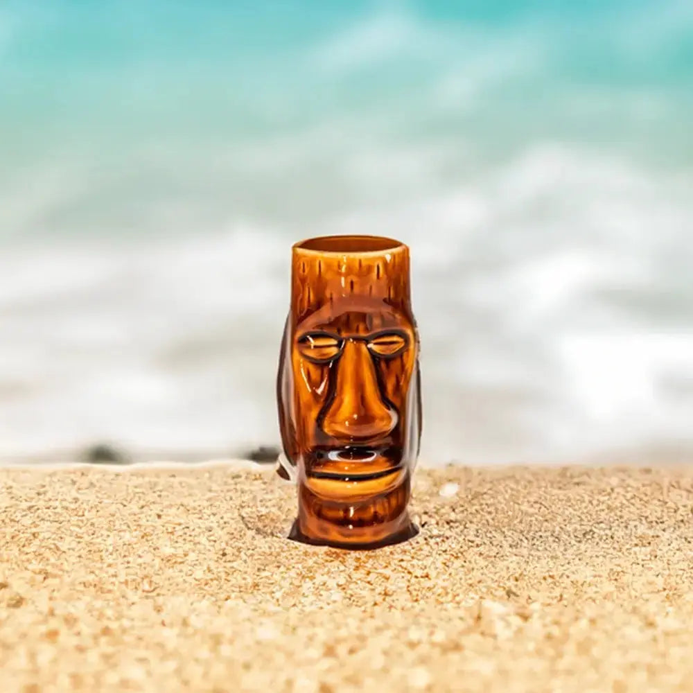 Verre tiki de style africain posé sur un plage de sable blanc. Au fond il y a la mer.