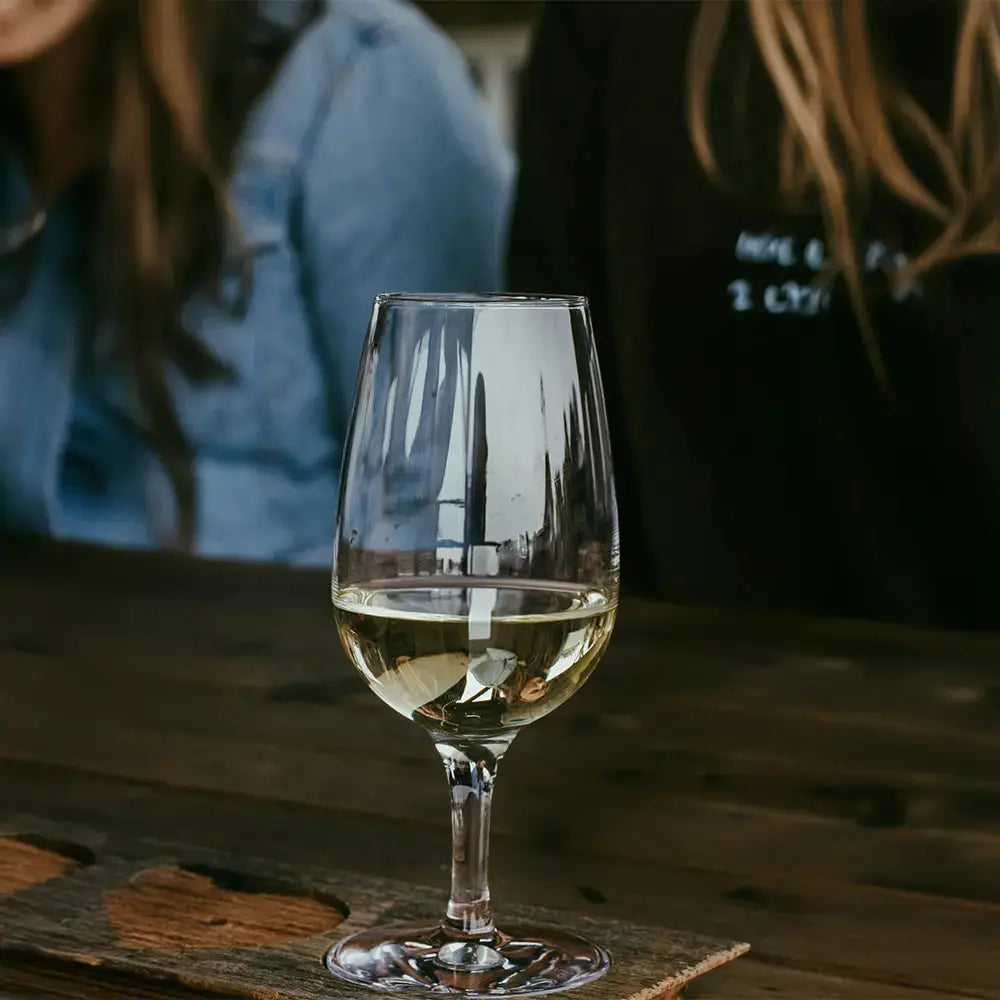 Verre inao universel rempli de vin blanc. Le verre est posé sur une table en bois. Il y a deux filles en arrière plan. 