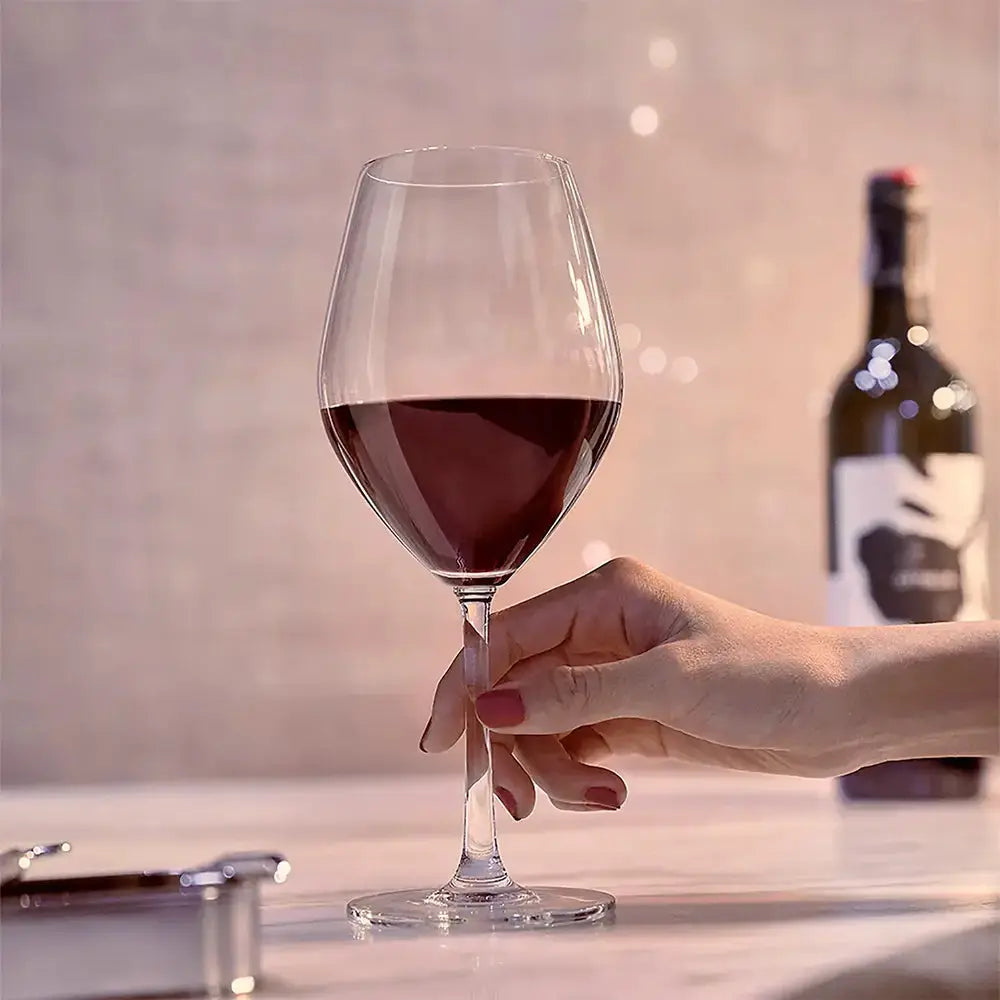 Verre pour vin rouge rempli de vin rouge. Une main de femme tiens le verre. Au fond de la photo il y a une bouteille de vin rouge.