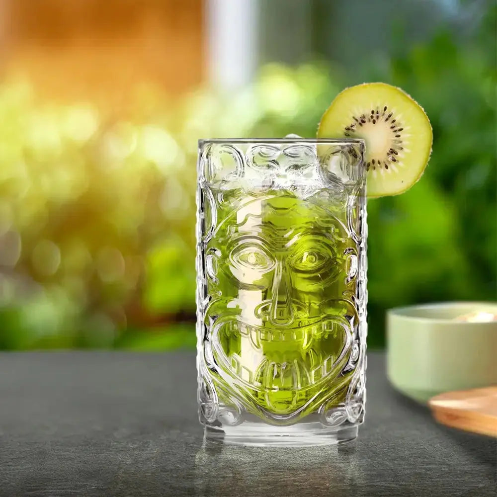 Verre Tiki transparent rempli d'un cocktail vert, décoré d'une tranche de kiwi. Présenté sur une table en extérieur avec un fond flou de verdure, ce verre sculpté au design tribal est idéal pour les cocktails exotiques et rafraîchissants.