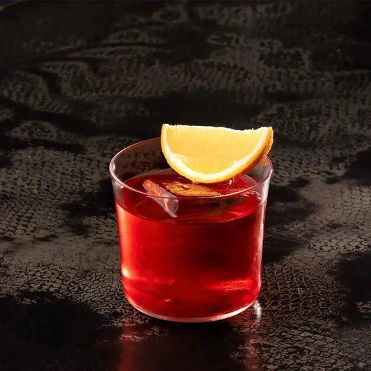 Verre à cocktail Lowball rempli d’un Negroni, surmonté d’une tranche d’orange, posé sur une table sombre texturée.