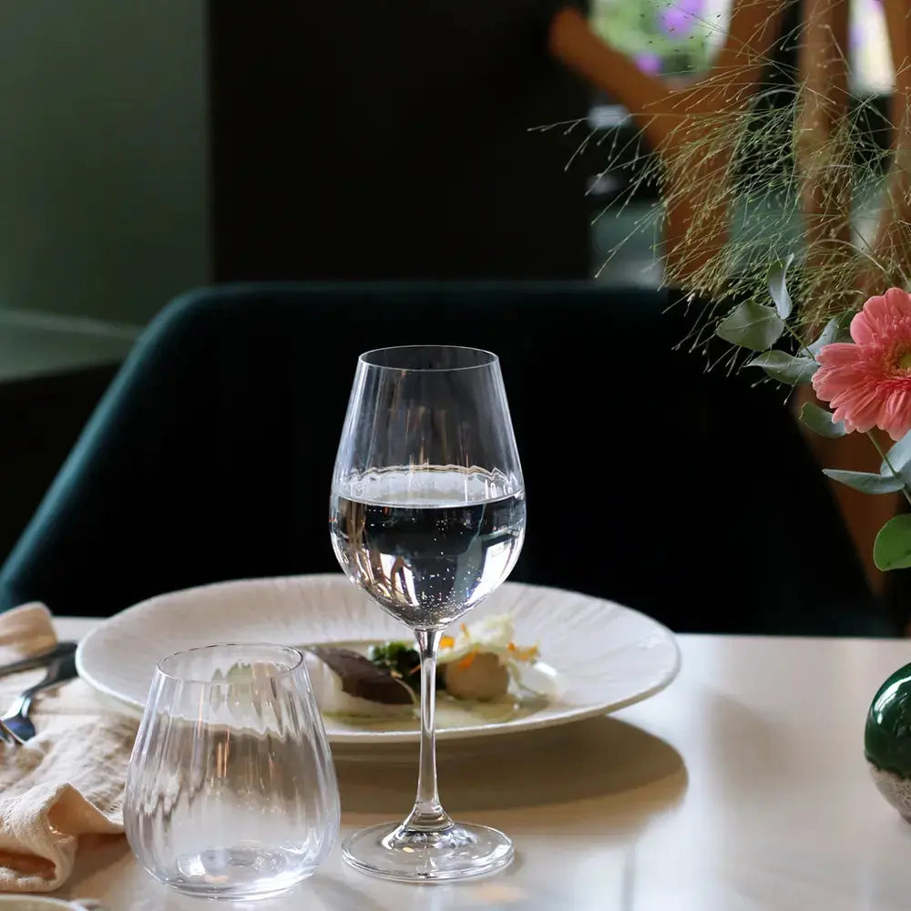 Verre à vin universel en cristal transparent, dressé sur une table élégante avec un décor floral et une assiette gastronomique en arrière-plan. Ambiance chaleureuse et raffinée, parfaite pour une dégustation.