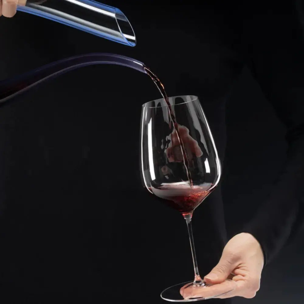 Une personne est en train de servir du vin depuis une carafe aux reflets bleus dans un verre à vin rouge original.