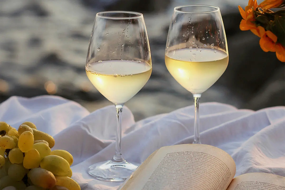 Deux verres de vin blanc sur une nappe blanche avec un livre ouvert à côté et une grappe de raisins blancs. 