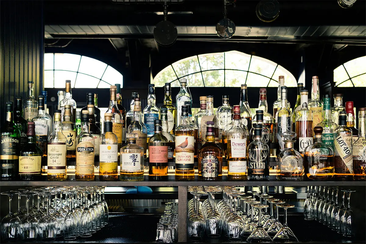 Derrière le comptoir d'un bar il y a des dizaines de bouteilles de spiritueux, certaines entamées, d'autres neuves. Devant, sous le bar il y a des verres pour déguster les spiritueux. 